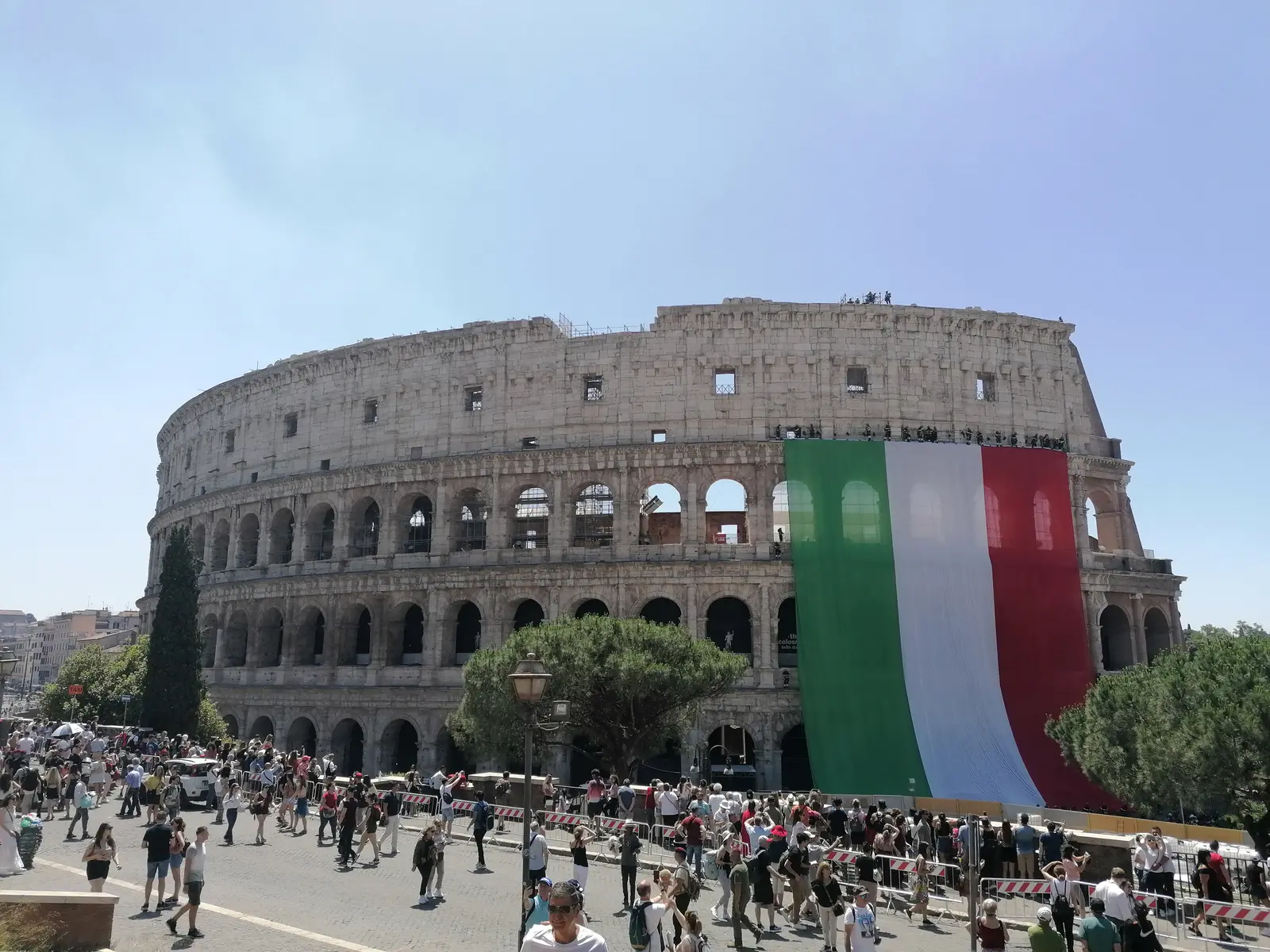The Colosseum in Rome with a giant Italian flag hanging from its side and crowds in front