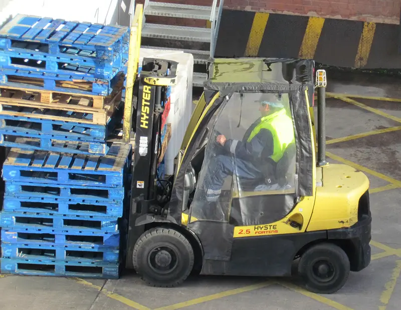 A forklift driver in a high-visibility vest moving next to stacked pallets