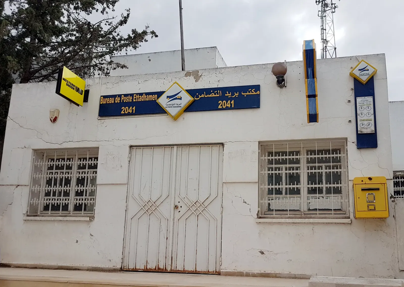 A white La Poste Tunisienne office in Ettadhamen with a blue sign in Arabic and French and a yellow letterbox